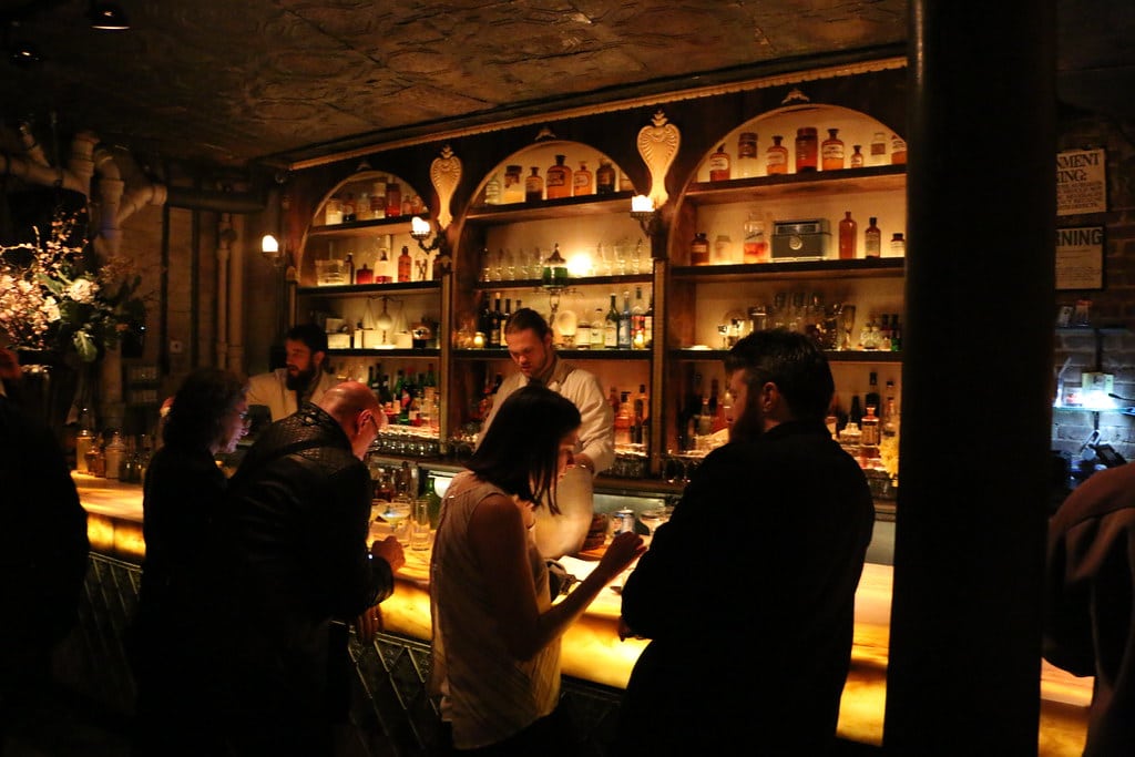 Bar bondé avec clients et barman, ambiance tamisée.